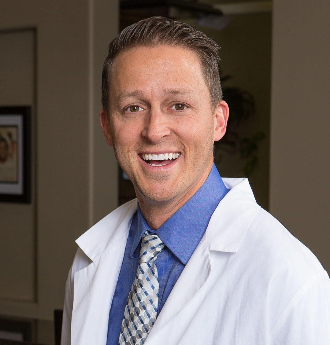A man in a white lab coat, blue dress shirt, and patterned tie stands in front of a softly lit background featuring a framed picture and green plants. His professional attire and setting suggest a clinical or healthcare-related environment.